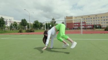 Uzun saçlı, eşofmanlı genç bir kadın sokak dansı yapıyor, dans salonu twerk 'i yapıyor, kalçalarını bir spor sahasında sallıyor. Şehirde serbest dans yapan bir şehir. Seks Dansı. Gimbal atışı.