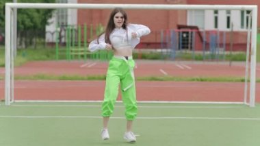 Uzun saçlı, eşofmanlı genç bir kadın sokak dansı yapıyor, dans salonu twerk 'i yapıyor, kalçalarını bir spor sahasında sallıyor. Şehirde serbest dans yapan bir şehir. Seks Dansı. Yavaş çekim