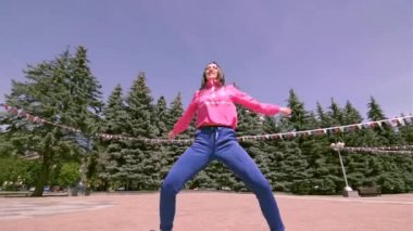 Eşofmanlı, uzun saçlı, enerjik genç bir kadın sokak dansı yapıyor. Şehir parkında açık bir alanda hip hop yapıyor. Gençlik alt kültürü. Çağdaş koreografi. Gimbal atışı.
