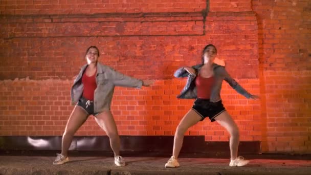 2人の美しい陽気な女性が現代の振付を踊り夜のレンガの壁に向かってストリートダンスをします 青年サブカルチャー — ストック動画