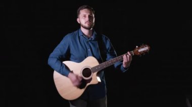 Orta boy, sakallı, akustik gitar çalan yakışıklı bir gitarist. Siyah arka plan. İzole edilmiş