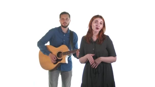 若い音楽家のデュエット 髭を生やした男がアコースティックギターを弾き 赤い髪の女性が歌う 白地だ — ストック動画