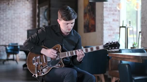 黒シャツのハンサムな男はカフェでギターの美しいブルースの組成物で遊んでいる レトロなスタジオ レンガのインテリア ジャズ ブルース ファンク 中ロングショット — ストック動画