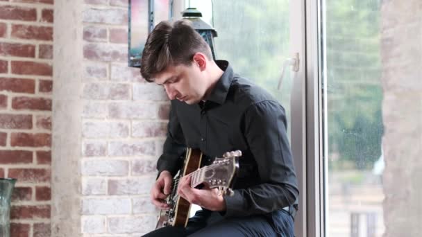 黒いシャツのハンサムな男が窓の向こうのカフェでギターを弾いてる 音楽の即興をブルース ジャズ ブルース ファンク 中ロングショット — ストック動画