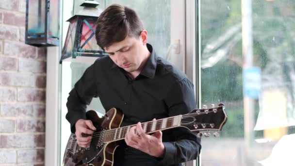 黒いシャツを着た才能あるギタリストが 窓の向こうのカフェでギターを弾き スイング ブルース ロック ロール ロック音楽を演奏する レトロなスタジオ レンガ造りのインテリア — ストック動画