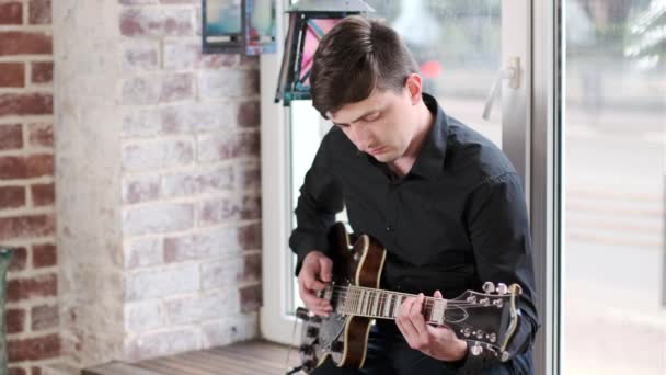 黒いシャツを着た才能あるギタリストが 窓の向こうのカフェでギターを弾き スイング ブルース ロック ロール ロック音楽を演奏する レトロなスタジオ レンガ造りのインテリア — ストック動画