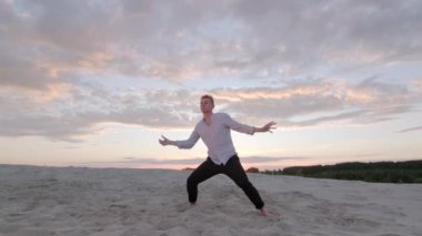 Yakışıklı dansçı gün batımında kumlu plajda serbest dans kompozisyonu sergiliyor. Çağdaş koreografi. Duygular. Yavaş çekim. Gimbal atışı.