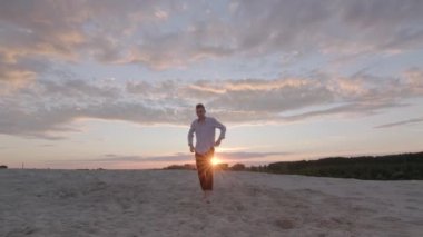 Genç düşünceli bir adam güzel bir yaz akşamında gün batımında kumda yalınayak yürüyor. Duygular. Yavaş çekim. Gimbal atışı.