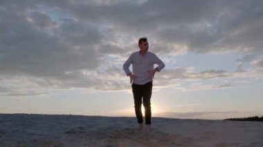 Yakışıklı dansçı gün batımında kumlu plajda serbest dans kompozisyonu sergiliyor. Çağdaş koreografi. Yavaş çekim. Canlı kamera hareket ediyor.