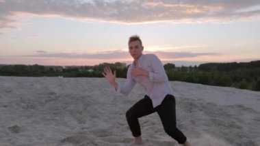 Şehvetli genç adam, bir yaz akşamı günbatımında, sık bir orman ve nehrin arka planına karşı kumda çağdaş koreografi yapar. Yavaş çekim. Canlı kamera hareket ediyor.
