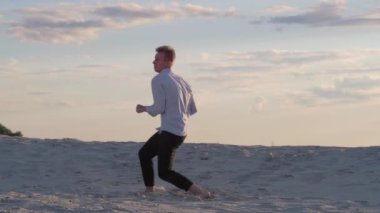 Şehvetli genç adam, güzel bir yaz akşamında gün batımında kumda çağdaş koreografi sergiliyor. Yavaş çekim görüntüleri