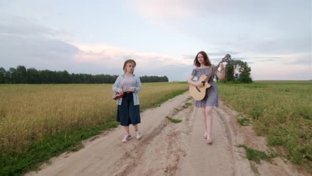 夏日的傍晚 两个年轻的女子沿着麦田边走着唱着 他们弹吉他和四弦琴 Gimbal Shot — 图库视频影像