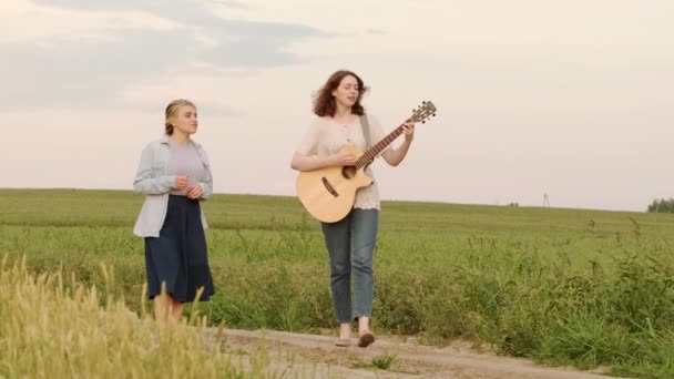 két gyönyörű nő sétál és énekel az út mentén egy búzamezőn egy nyári estén. A lány elkíséri a gitárt. Lassított felvétel