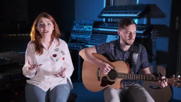 一个留着胡子的英俊吉他手和一个红头发的女孩一起坐在音乐工作室的吉他上 手控摄像头中长枪 — 图库视频影像