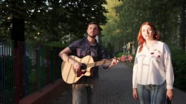 Sakallı bir adam akustik gitar çalar, kızıl saçlı bir kadın bir yaz akşamı gün batımında şehir parkında şarkı söyler.