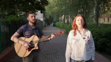 Genç müzisyenlerin düeti. Adam gitar çalıyor. Kadın şarkı söylüyor. Şehir parkında bir yaz akşamı gün batımında