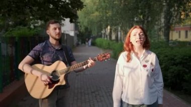 Genç müzisyenlerin düeti. Adam gitar çalıyor. Kadın şarkı söylüyor. Şehir parkında bir yaz akşamı gün batımında
