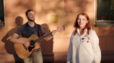 Sakallı yakışıklı gitarist gitar çalıyor, kızıl saçlı kız yaz akşamı gün batımında dışarıda şarkı söylüyor.