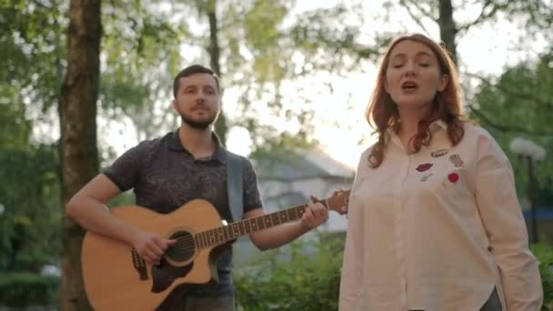 一个留着胡子的男人会弹吉他 一个红头发的女人会在夏夜黄昏的公园里唱歌 — 图库视频影像
