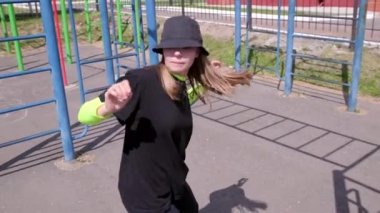 Sportsground 'da eşofmanlı ve şapka dansı yapan neşeli genç bir kadın. Şehirde serbest dans yapan bir şehir. Gençlik alt kültürü. Yavaş çekim