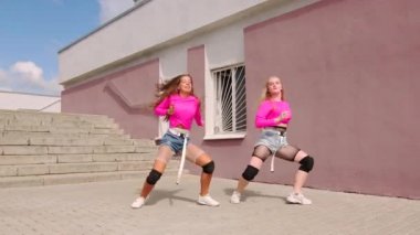 Kısa şortlu, serbest stil sokak dansı yapan, merdivenli bir binanın önünde hip hop koreografisi yapan neşeli genç kızların düeti. Yavaş çekim