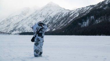 Kış, kamuflaj elbiseli bir adam dağların ortasında karla kaplı bir göl üzerinde duruyor ve dürbünle bakıyor.