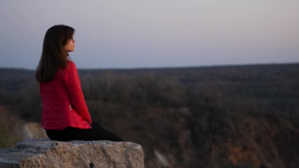 穿着红色夹克的年轻女孩坐在岩石上 望着远处 — 图库视频影像