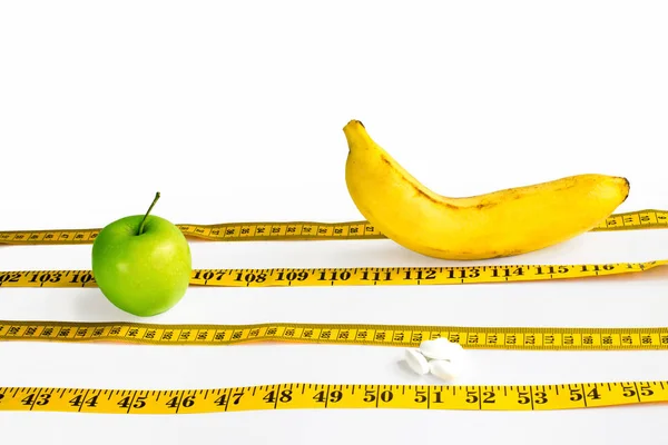 Gelbes Zentimeterband als Sportbahnen angelegt, auf denen grüne Äpfel, Schalen und Bananen auf weißem Hintergrund miteinander konkurrieren — Stockfoto