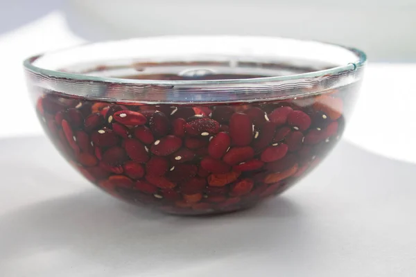 Beaucoup de haricots rouges crus trempés dans l'eau dans un bol en verre clair sur une table blanche avec parasol Photo De Stock