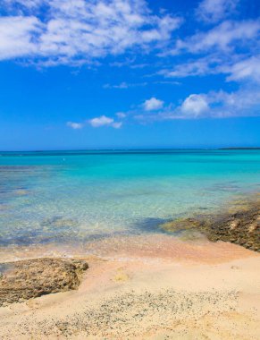 Pembe kumları, taşları ve turkuaz suları ve mavi gökyüzünde bulutları olan plaj..