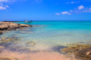 Pembe kumları, taşları ve turkuaz suları ve mavi gökyüzünde bulutları olan plaj..