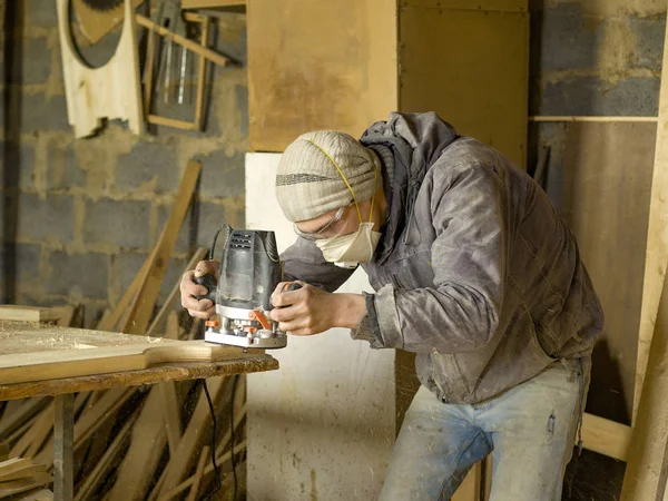 Человек обрабатывает дерево в деревообрабатывающей мастерской, плотник в респираторе с фрезерным станком — стоковое фото