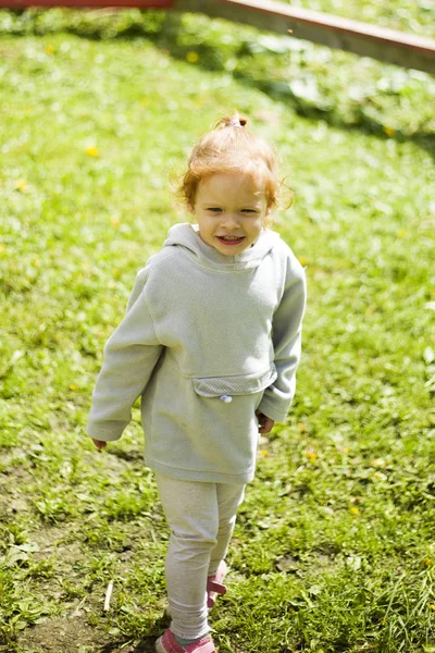 Маленькая счастливая рыжая девочка бегает по подстриженной зеленой траве в детском парке под приятным солнцем — стоковое фото