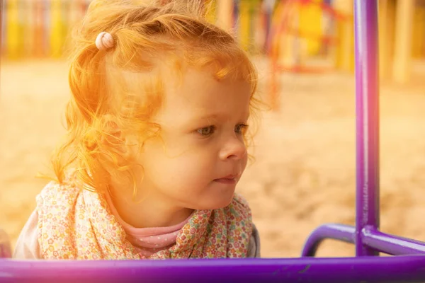 लाल बालों वाली छोटी लड़की खेल के मैदान पर एक बच्चों के पार्क में खेलती है और कैमरा लेंस सूर्य से ऑरेंज सनसेट लाइट्स उठाती है — स्टॉक फ़ोटो, इमेज