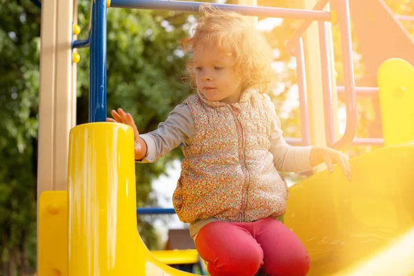 Маленькая рыжая девочка сидит на холме в детском парке и греется под теплым солнцем — стоковое фото