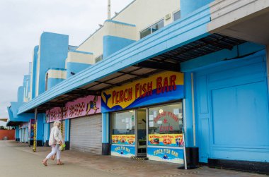 New Brighton, İngiltere: 3 Haziran 2020: Bir sokak görüntüsü, Corona virüsünün geçici olarak kapanması için gerekli olan işletmeler üzerindeki etkisini gösteriyor. Yalnız bir kadın açık olan levrek lokantasının önünden geçiyor.