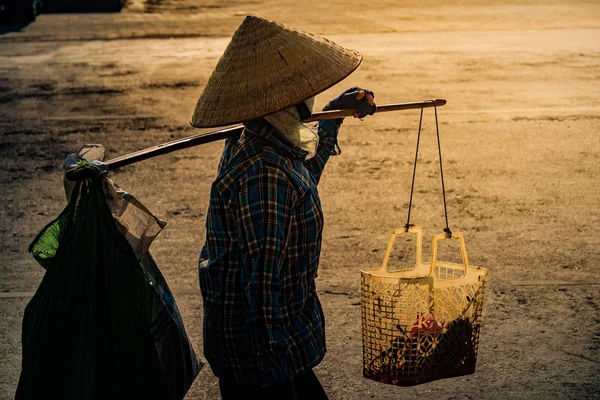 Vietnamese with conical hat carries a yoke on her shoulder along the street.