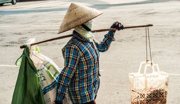 Vietnamese with conical hat carries a yoke on her shoulder along the street.