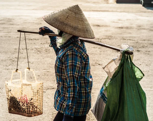 Vietnami kúpos kalapban egy igát hordozza a vállán az utcán. — Stock Fotó
