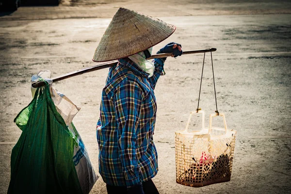 Vietnamesin mit kegelförmigem Hut trägt auf der Straße ein Joch auf der Schulter. — Stockfoto