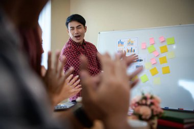 Handsome young man standing near whiteboard and pointing on the chart presentation perfect planning business with people group clapping.