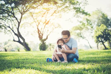 Baba ve kızı parkta akıllı telefon kullanarak gülümsüyor.
