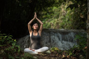 Yoga yapan genç bir kadın şelalenin yanında oturuyor.
