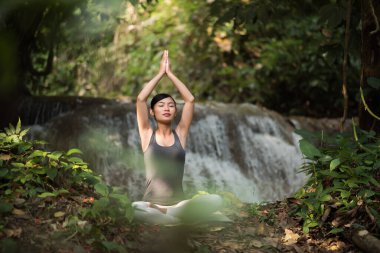 Yoga yapan genç bir kadın şelalenin yanında oturuyor.