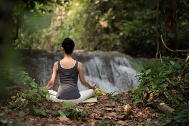 Yoga yapan genç bir kadın şelalenin yanında oturuyor.