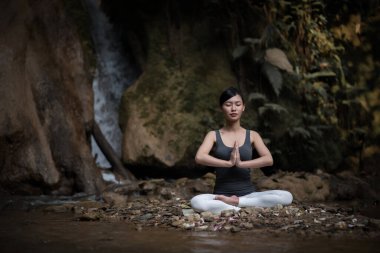 Yoga yapan genç bir kadın şelalenin yanında oturuyor.