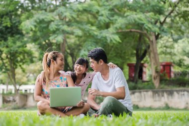 Mutlu aile babası, anne ve kızı çimlerin üzerinde oturup dışarıda dizüstü bilgisayar oynuyorlar.