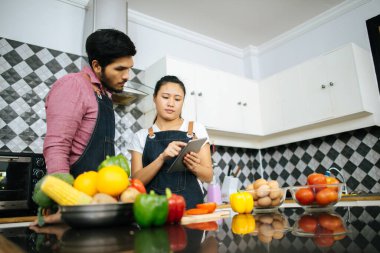 Mutlu genç çift, evde yemek pişirirken dijital tablet kullanarak yemek yapma yöntemini kullanıyor..