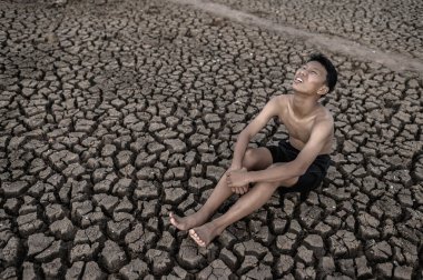 Çocuk dizlerini bükerek oturur ve kuru toprakta yağmur istemek için gökyüzüne bakar..