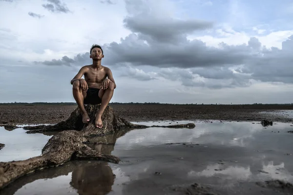 A man sat bent his knees, looking at the sky at the base of the tree and surrounded by water.